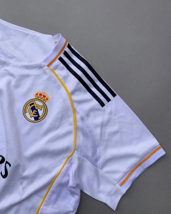 Real Madrid 2025/26 home jersey with classic white design, black adidas stripes, golden-yellow trim, Emirates Fly Better sponsor, and embroidered club crest – official fan and player kit